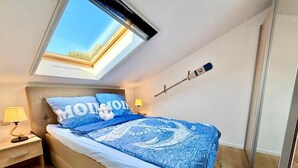 Ein Schlafzimmer mit Dachfenster, einem Bett mit blauer Bettwäsche, einer Nachttischlampe und einem an der Wand befestigten Thermometer.