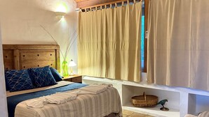 Una camera da letto con un letto, un comodino, una lampada, una finestra con tende e un piccolo ripiano con un cesto e una statuetta di pappagallo.