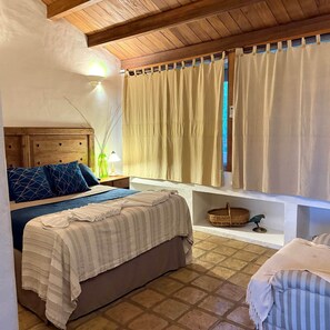 Un dormitorio con cama, mesita de noche, lámpara, ventana con cortinas y una pequeña repisa con una cesta y una figurita de loro.
