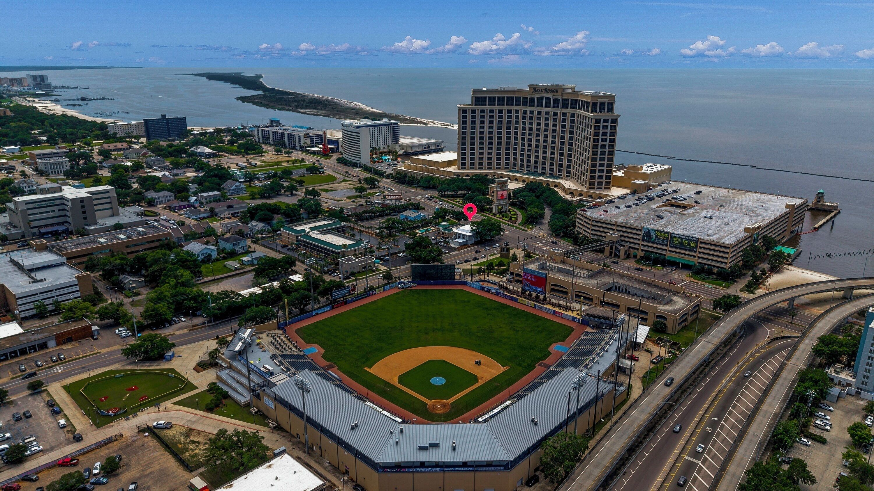 An aerial view of a baseball stadium with a parking lot, a large building, and a body of water in the background.