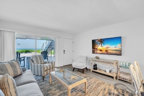 Ein Wohnzimmer mit Sofa, Sesseln, Couchtisch, Fernseher und Blick auf den Strand.