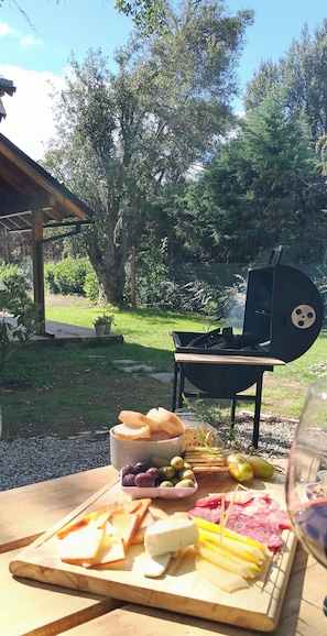 An outdoor dining area with a wooden table set for a meal, a grill in the background, and a garden view.