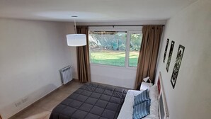 A bedroom with a bed, a bedside table, a lamp, a window with curtains, and a radiator. - Between Lakes and Mountains • Your Place in Bariloche (San Carlos de Bariloche)