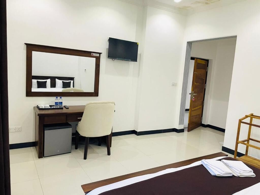 A hotel room with a bed, a desk with a chair, a small refrigerator, a television mounted on the wall, and a wooden door.