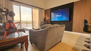 A hotel room with a large flat-screen TV displaying the Disney+ logo, a grey sofa, a wooden coffee table, and a balcony with a view.