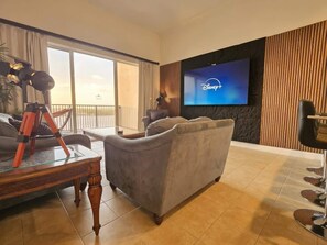 A hotel room with a large flat-screen TV displaying the Disney+ logo, a grey sofa, a wooden coffee table, and a balcony with a view. - Lakefront Condo 1 Mile to Disney w/ Pool, Hot Tub (Orlando)