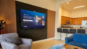 A living room with a flat-screen TV displaying Disney+ Hulu, a grey armchair, a blue sofa, and a coffee table with rolled towels. - Lakefront Condo 1 Mile to Disney w/ Pool, Hot Tub (Orlando)