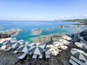 Ein Strand mit klarem, blauem Wasser, Felsformationen und Liegestühlen mit Sonnenschirmen.