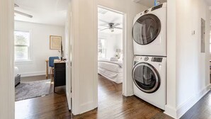 Ein Waschraum mit zwei nebeneinander gestapelten Frontlader-Waschmaschinen und -Trocknern, einem Deckenventilator und einem Bett im angrenzenden Schlafzimmer.
