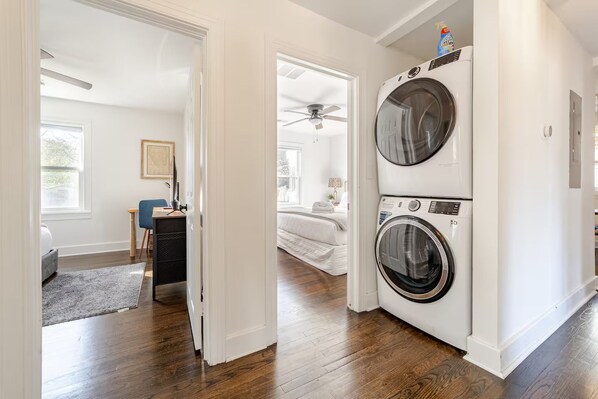Uma lavanderia com duas máquinas de lavar e secar empilhadas, um ventilador de teto e uma cama no quarto adjacente.