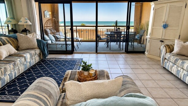 A living room with a sofa, a coffee table with a plant, and a view of the beach through large glass doors. - Oceanfront Condo with Heated Pool, 2 Balconies, Beach Access & Courts (Ocean City)