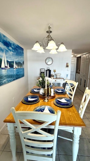 Une salle à manger avec une table en bois, des chaises blanches, un lustre et un tableau représentant des voiliers.