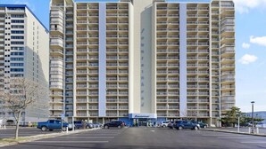 A multi-story hotel with a central vertical feature, a parking lot in front, and a clear sky. - Oceanfront Condo with Heated Pool, 2 Balconies, Beach Access & Courts (Ocean City)