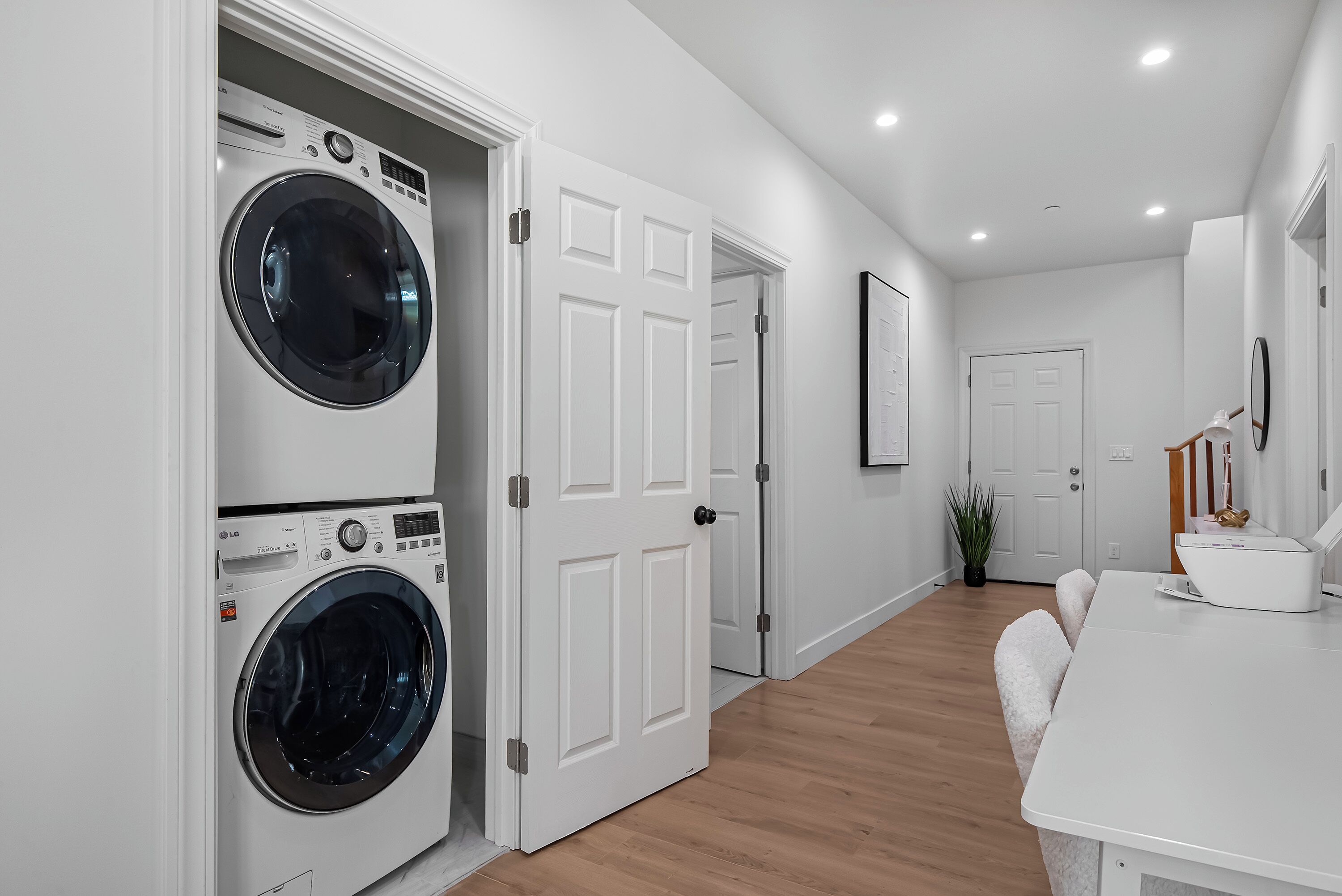 A laundry room with two stacked front-loading washing machines, a white door, a hallway leading to a room with a white table and a plant.