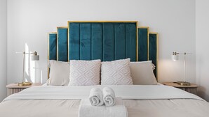 Cama bem arrumada com lençóis brancos e toalhas enroladas, cabeceira estofada em tom de azul-esverdeado e duas mesas de cabeceira com abajures.