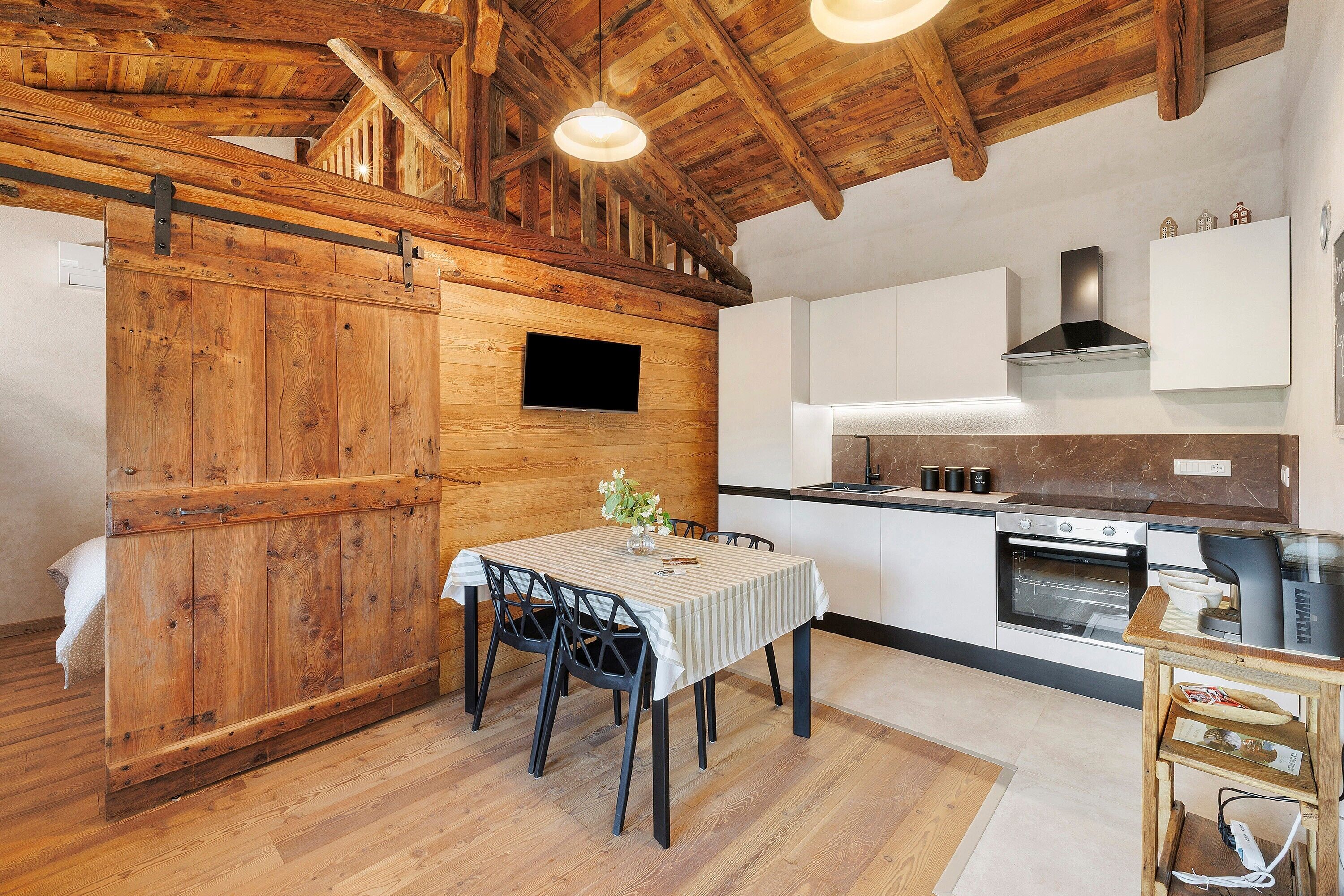 A rustic kitchen with wooden beams, a dining table with chairs, a TV mounted on the wall, and modern appliances.