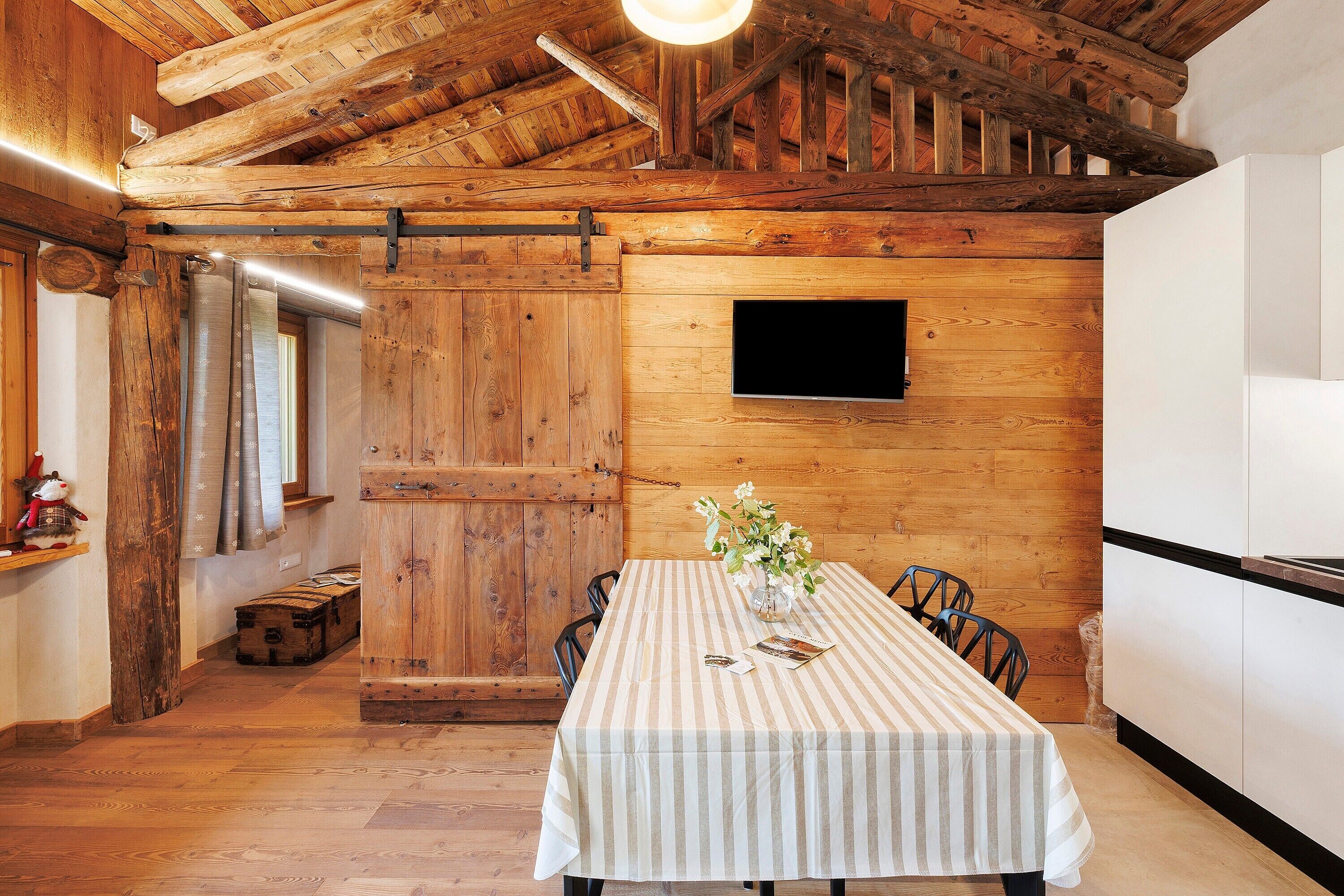 Un interno rustico in legno con un tavolo da pranzo, sedie, un vaso con fiori, un baule di legno e una televisione a parete.