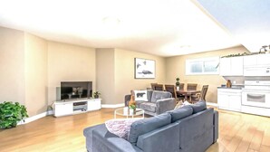 Una sala moderna con un sofá gris, un televisor, un comedor con sillas y una cocina con gabinetes blancos.