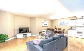 Ein modernes Wohnzimmer mit grauem Sofa, Fernseher, Essbereich mit Stühlen und einer Küche mit weißen Schränken.