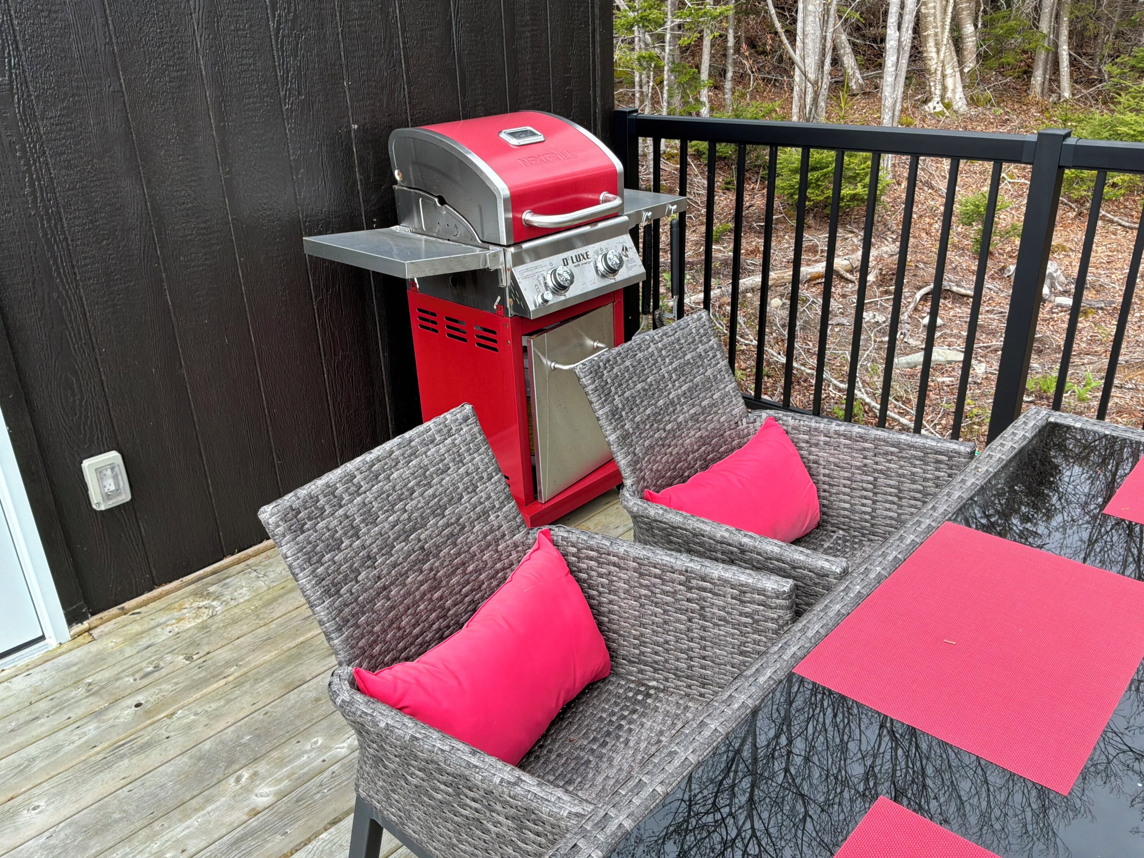 A patio with wicker chairs and red cushions, a red grill, and a black fence.