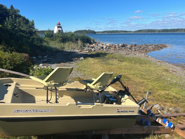 A boat named 'SPORTSMAN8' with two chairs and a fishing rod, docked near a rocky shore with a lighthouse in the background.