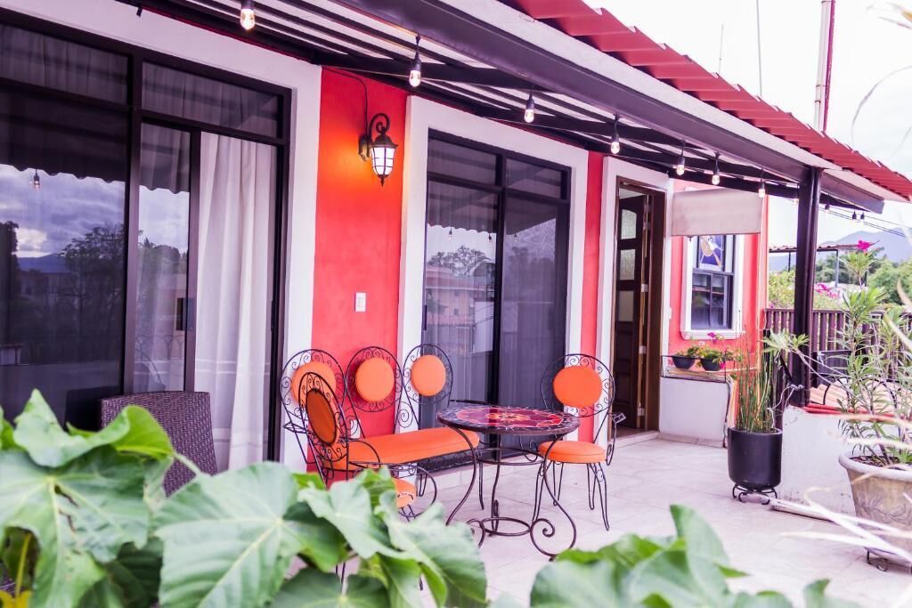 Un patio con mobiliario exterior, una planta en maceta y un edificio de fachada roja y grandes ventanales.