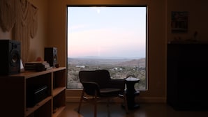 Una habitación con un ventanal grande, un escritorio de madera, una silla, una estantería y un altavoz.