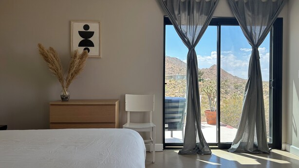 Un dormitorio con cama, cómoda, silla, ventana con cortinas y vistas a un paisaje desértico.