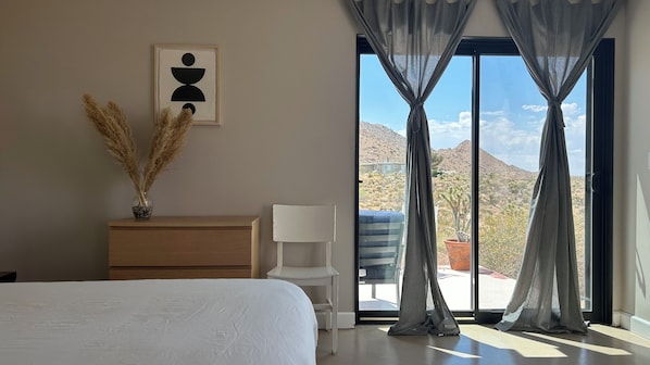 Ein Schlafzimmer mit Bett, Schminktisch, Stuhl, einem Fenster mit Vorhängen und Blick auf eine Wüstenlandschaft.