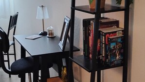A dining area with a table, chairs, a lamp, a bookshelf with books and decorative items, and a potted plant.