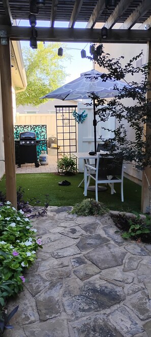 Un espace patio avec un sol en pierre, une table et une chaise blanches, un barbecue et un parasol bleu.