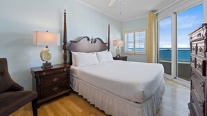 A hotel room with a large bed, a wooden nightstand, a dresser, a chair, and a view of the ocean through a window. - Beautiful Beach Front Condo with Breathtaking Views (Orange Beach)