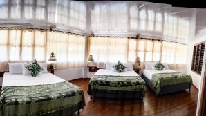 Habitación de hotel con cuatro camas dispuestas en una esquina, ventanas amplias con cortinas translúcidas y suelo de madera.