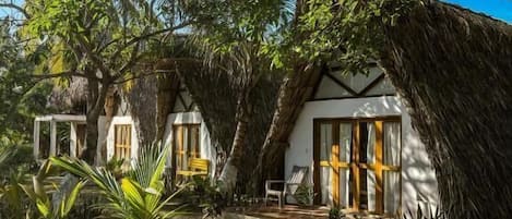 A thatched-roof bungalow with a stone pathway leading to the entrance, surrounded by lush greenery and a clear blue sky.