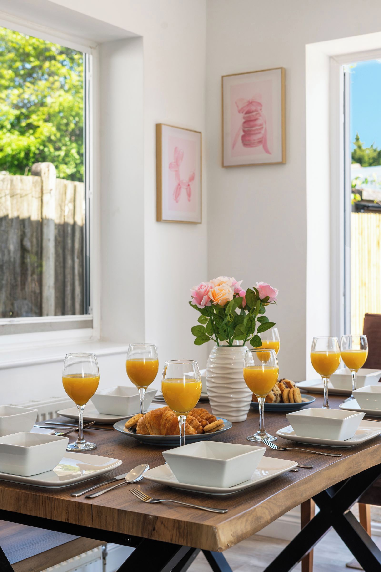 Una mesa de desayuno dispuesta con cruasanes, jugo de naranja y un florero con flores.