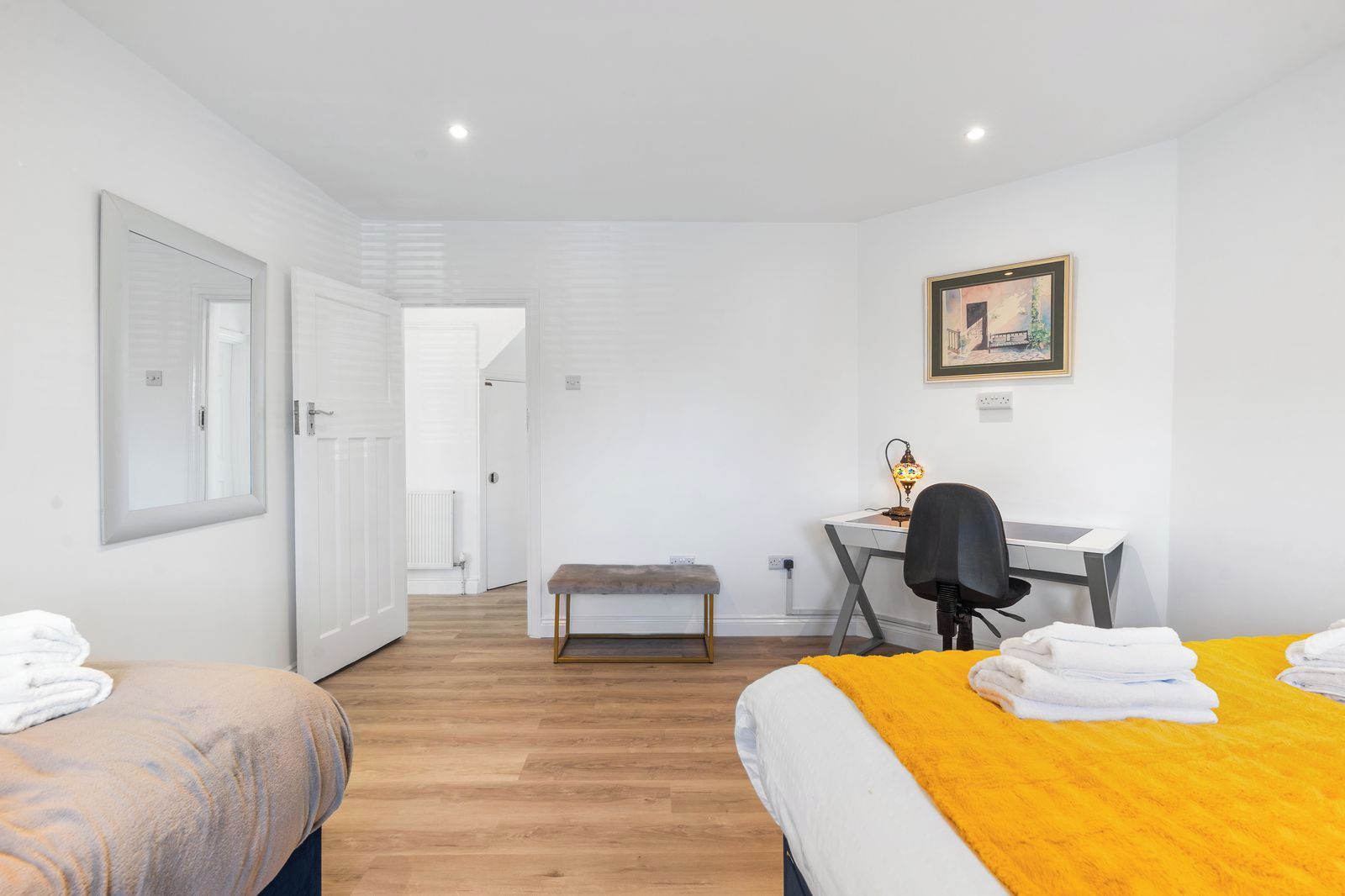 A room with two beds, a desk with a chair, a bench, and a painting on the wall.