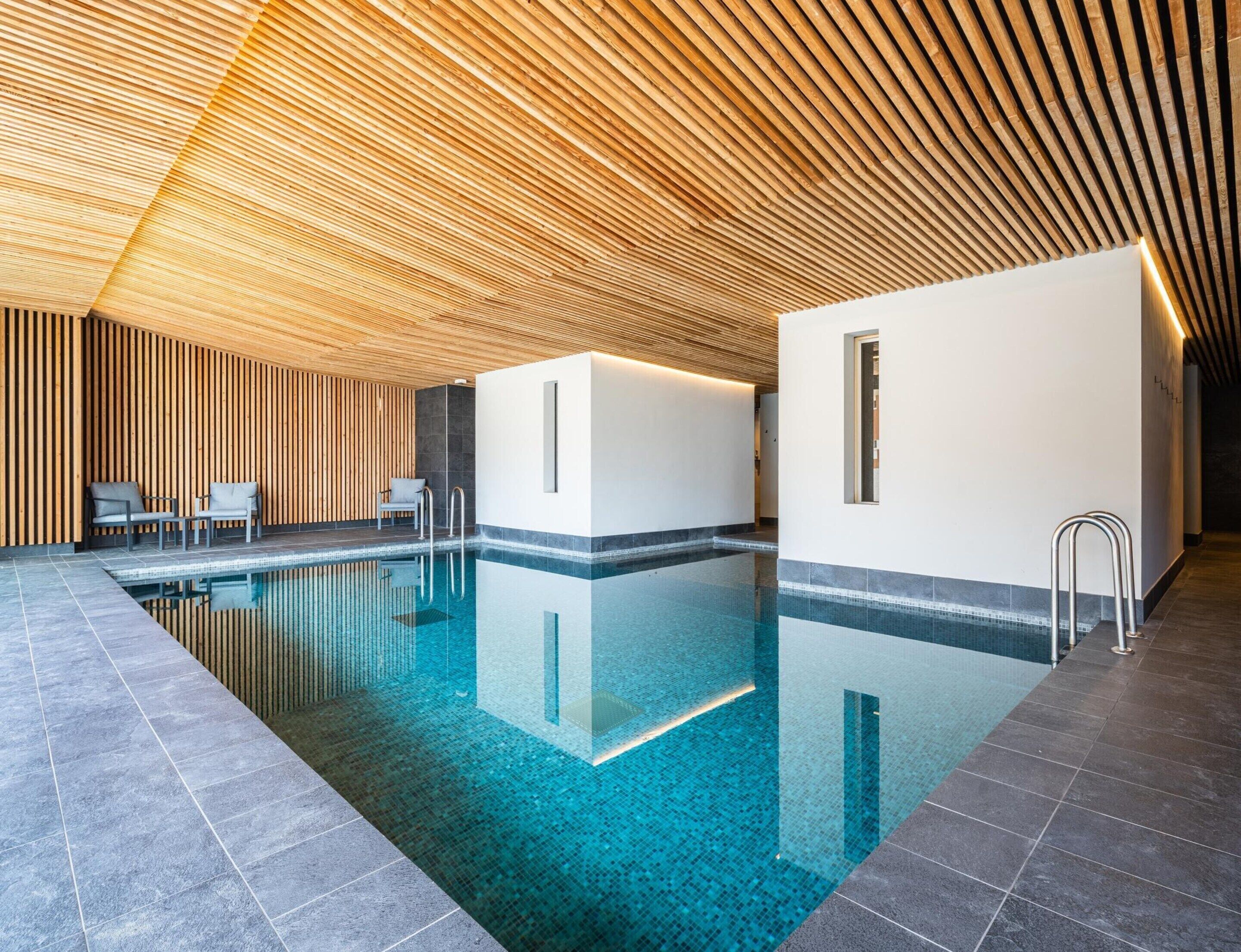 Une piscine intérieure moderne avec un plafond en lambris de bois, une eau cristalline et un sol carrelé.