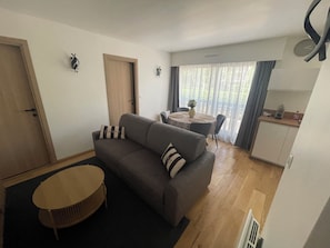 A living room with a sofa, a round coffee table, a dining table with chairs, a kitchen area, and a door. - Cosy apartment 300 metres from the beach (Deauville)