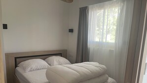 Una cama bien hecha con cabecera de madera, una ventana con cortinas translúcidas y un interruptor de luz en la pared.