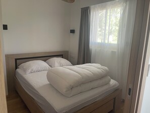 A neatly made bed with a wooden headboard, a window with sheer curtains, and a light switch on the wall. - Cosy apartment 300 metres from the beach (Deauville)