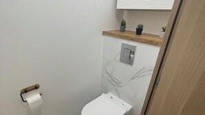 Un baño pequeño con inodoro, piso de mármol, estante de madera con plantas y techo blanco.