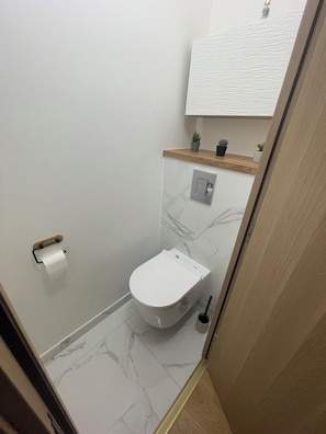 A small bathroom with a toilet, a marble floor, a wooden shelf with plants, and a white ceiling. - Cosy apartment 300 metres from the beach (Deauville)