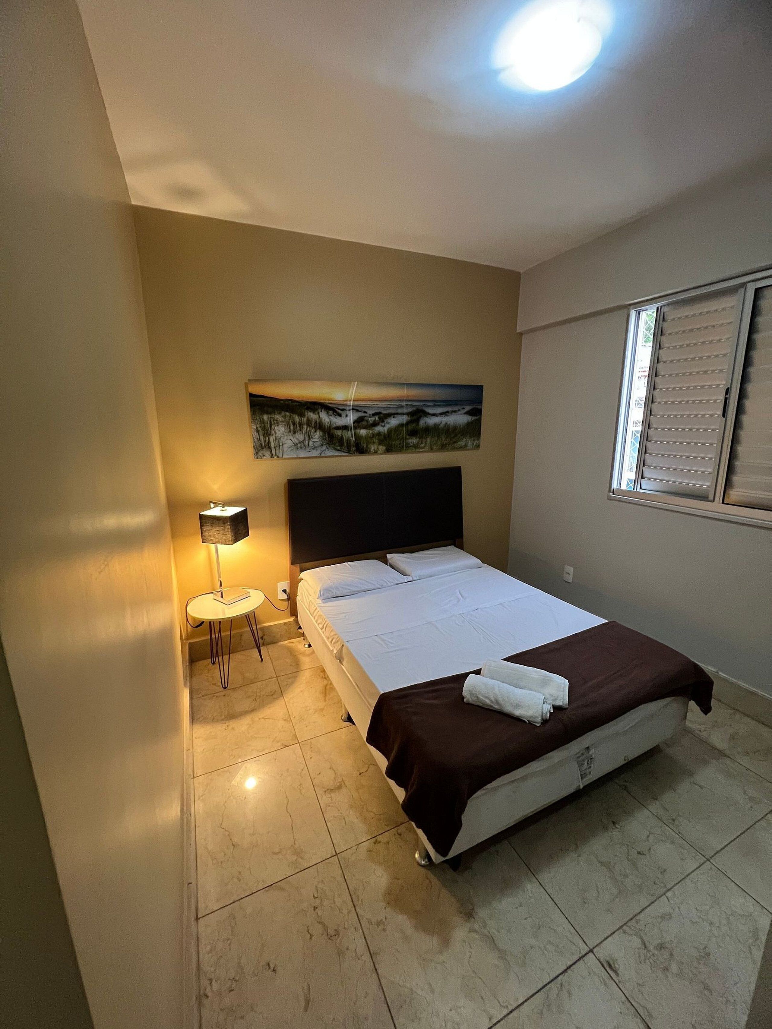 A hotel room with a bed, a painting on the wall, a window with blinds, a small table with a lamp, and a tiled floor.