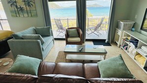 A living room with a sofa, armchairs, a coffee table, and a TV stand. A balcony with a view of the sea and mountains is visible through large windows. - NEW LISTING: Stylish, scenic & renovated condo steps from Caribbean in Red Hook! (St Thomas)