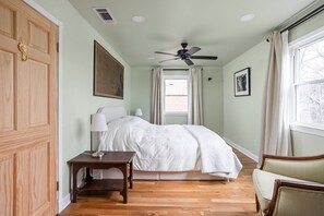 Ein Schlafzimmer mit einem Bett, einem Nachttisch, einem Sessel, einem Deckenventilator und einem Fenster mit Vorhängen.