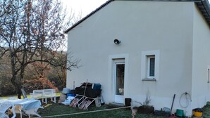 Un edificio bianco con una porta, finestre, una panchina e vari oggetti nel giardino, come un tubo da giardinaggio e una pianta in vaso.