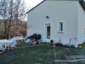 Ein weißes Gebäude mit einer Tür und Fenstern, einer Bank und verschiedenen Gegenständen wie einem Schlauch und einem Blumentopf im Garten.