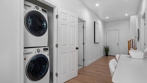 Une buanderie avec deux machines à laver superposées, une porte blanche, un couloir, un coin repas avec une table et des chaises, et une plante en pot.