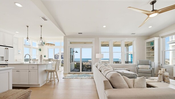 A modern kitchen with white cabinetry, a dining area with a view, a living room with a beige sofa, and a ceiling fan. - Ocean Views! One Row to the Beach with Pool & Spa (Oak Island)
