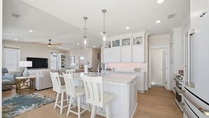 A modern kitchen with white cabinetry, a central island with bar stools, and a dining area with a sofa and coffee table. - Ocean Views! One Row to the Beach with Pool & Spa (Oak Island)